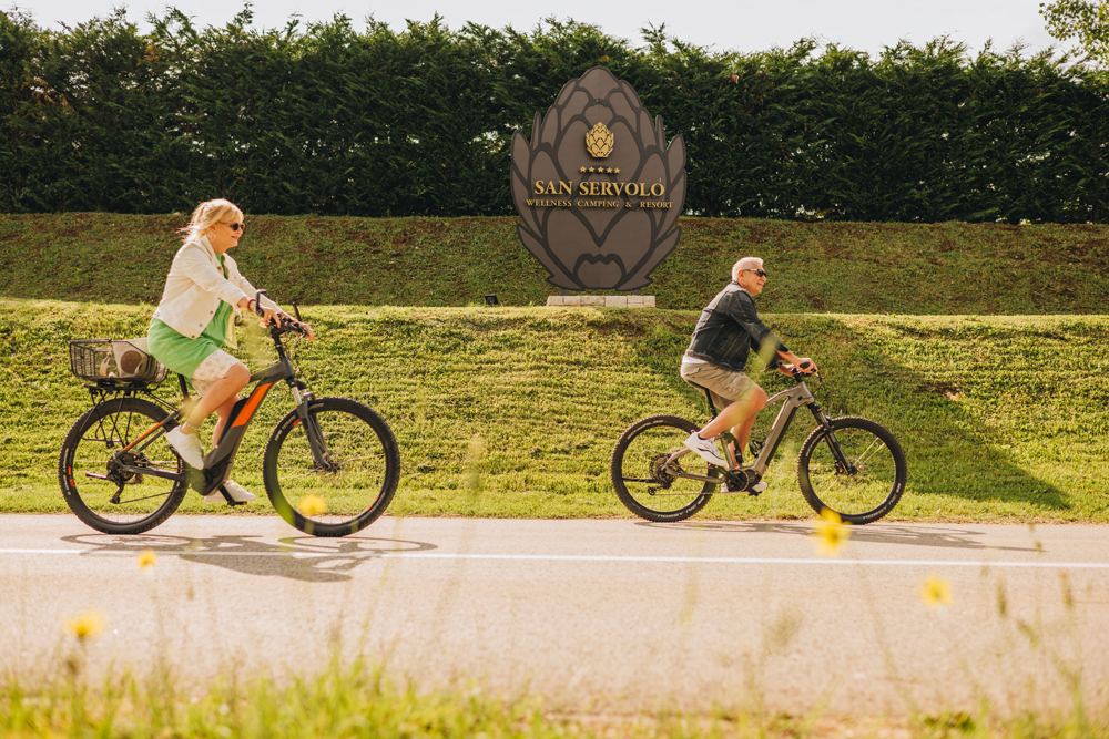 Zwei ältere Personen beim Radfahren mit E-Bikes vor dem Schild des San Servolo Camping Resorts in Kroatien.