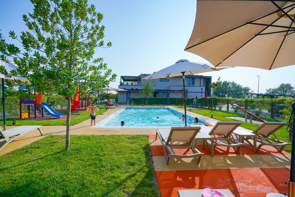 Swimmingpool im Resort San Servolo mit Liegestühlen und Spielgeräte für Kinder links und rechts vom Pool sowie
Haus im Hintergrund.