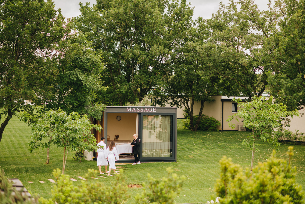 Massage-Angebot im San Servolo Resort. Auf dem Bild ist ein Paar in Bademänteln zu sehen, welches
von einer Resort-Mitarbeiterin in ein kleines Haus mitten im Grünen eingeladen wird, auf dem Massage über dem Eingang steht.