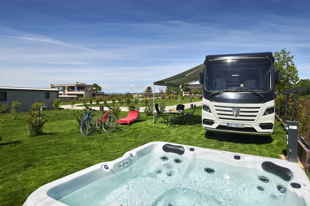 Premium Luxury Stellplatz auf dem Resort San Servolo mit privatem Whirlpool. Auf dem Platz
steht ein Wohnmobil mit Vorzelt. Unter dem Vorzelt ein Tisch mit 4 Stühlen sowie zwei Fahrräder daneben.