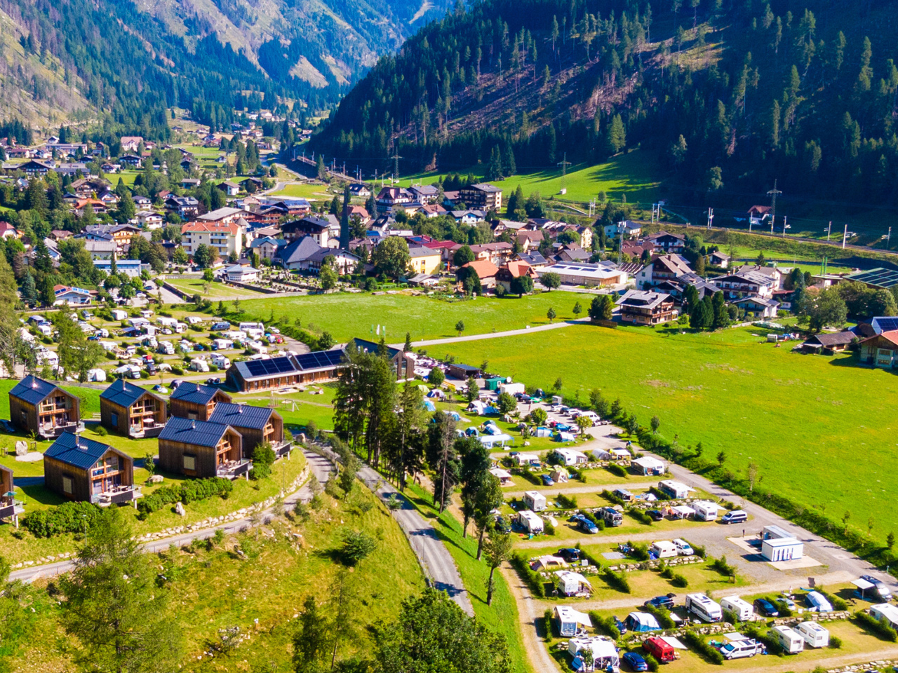 Luftaufnahme des Camping HOCHoben.
                           Man sieht die Stellplätze belegt mit Zelten, Wohnwägen und Wohnmobilen, sowie Chalets und im Hintergrund das Dorf Mallnitz
                           in Kärnten.