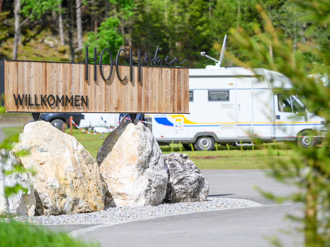 Einfahrt zum Camping HOCHoben mit Willkommensschild und Camper, der auf
                           einem Stellplatz steht im HIntergrund.